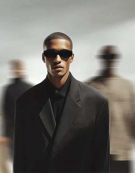 Man in black oversized blazer and dark sunglasses stands with two blurred figures behind him.