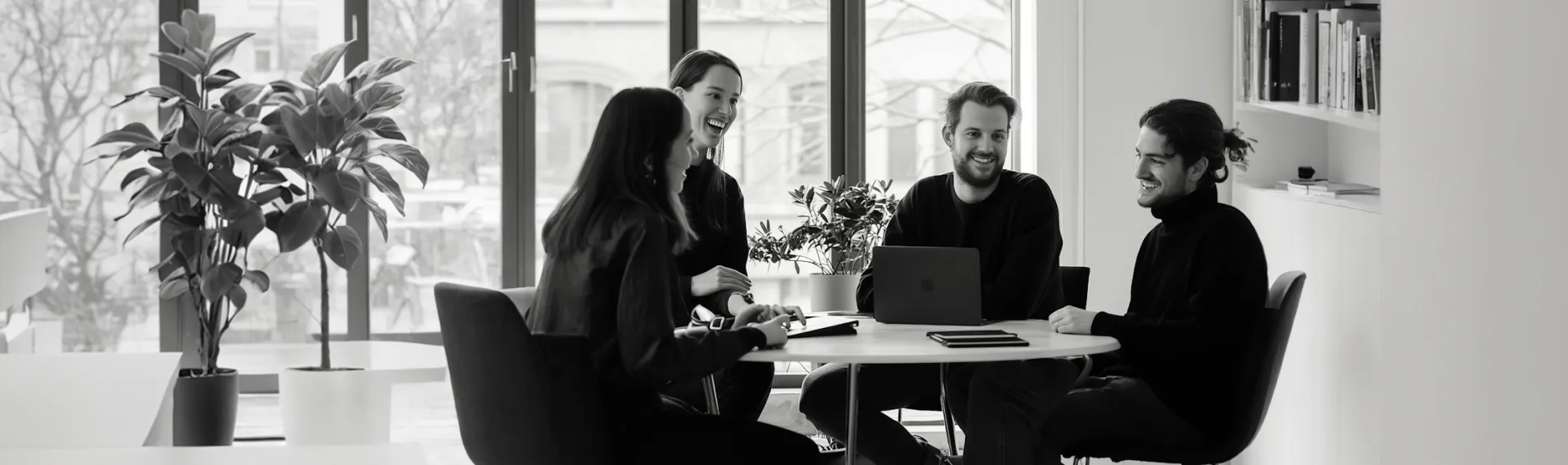 Four young professionals smiling and talking around a table with a laptop in a bright modern office.