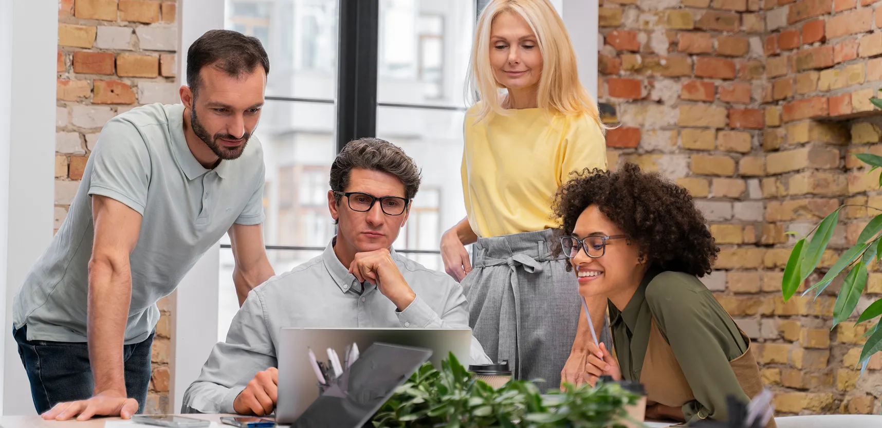 Four colleagues collaborate around a laptop, focused in a modern office with brick walls and plants.