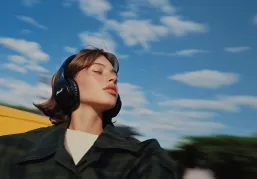 Woman with short hair wearing headphones and a dark coat against a blue sky with clouds.