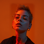 Person with short hair, large round glasses, and hoop earrings lit by warm orange light.