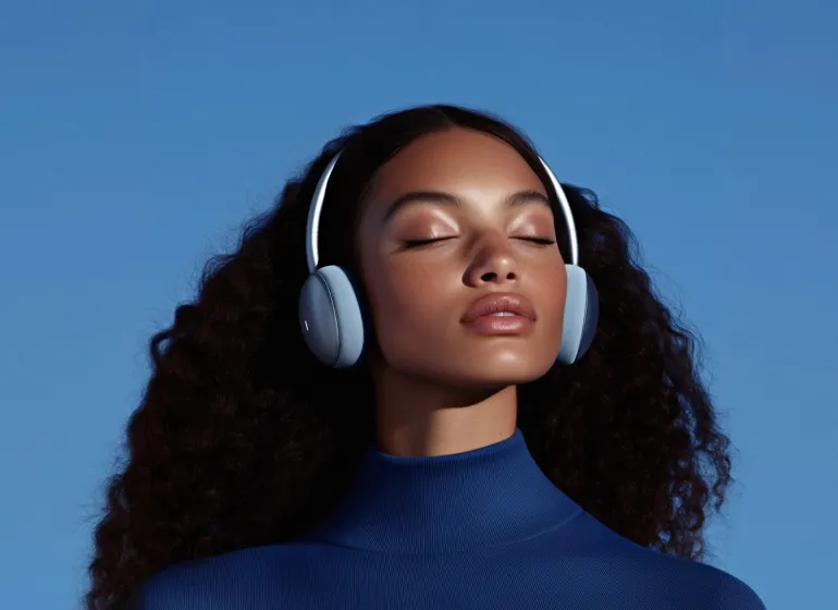 Woman with curly hair wearing headphones and a blue turtleneck with closed eyes against blue background.