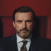 Portrait of a man with a beard and mustache wearing a suit and tie against a dark red background.