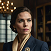 Woman with short brown hair wearing a dark blazer, looking to the side against a blue background.