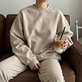 Person in beige sweatshirt and pants holding a cup of iced coffee, sitting on a brown couch.