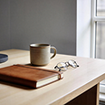 Coffee mug next to eyeglasses and a closed wooden notebook on a light wood table by a window.