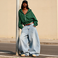 Person in oversized light blue jeans and green off-shoulder top standing with one hand in pocket.