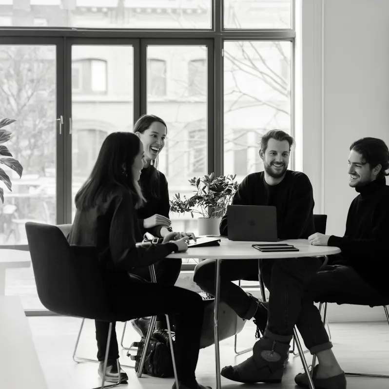 Four people smiling and chatting around a table in a bright office with large windows.