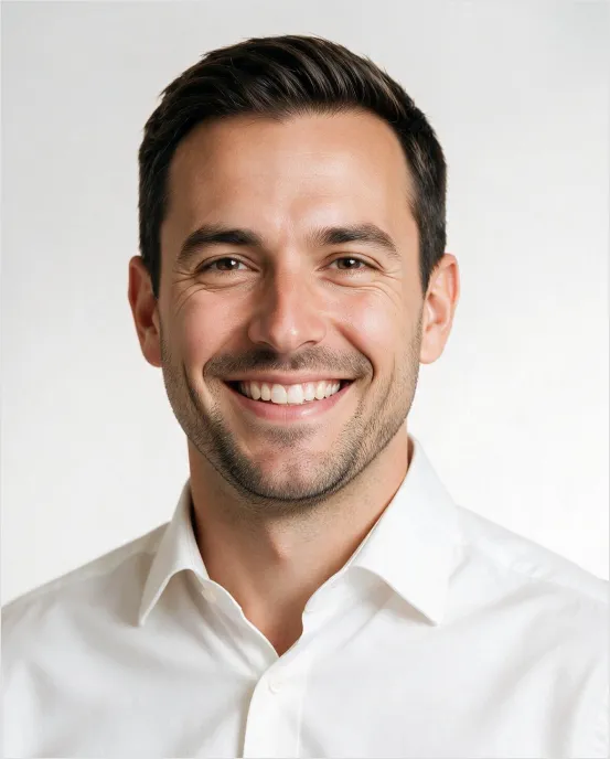 Smiling man with dark hair wearing a white collared shirt against a plain background.