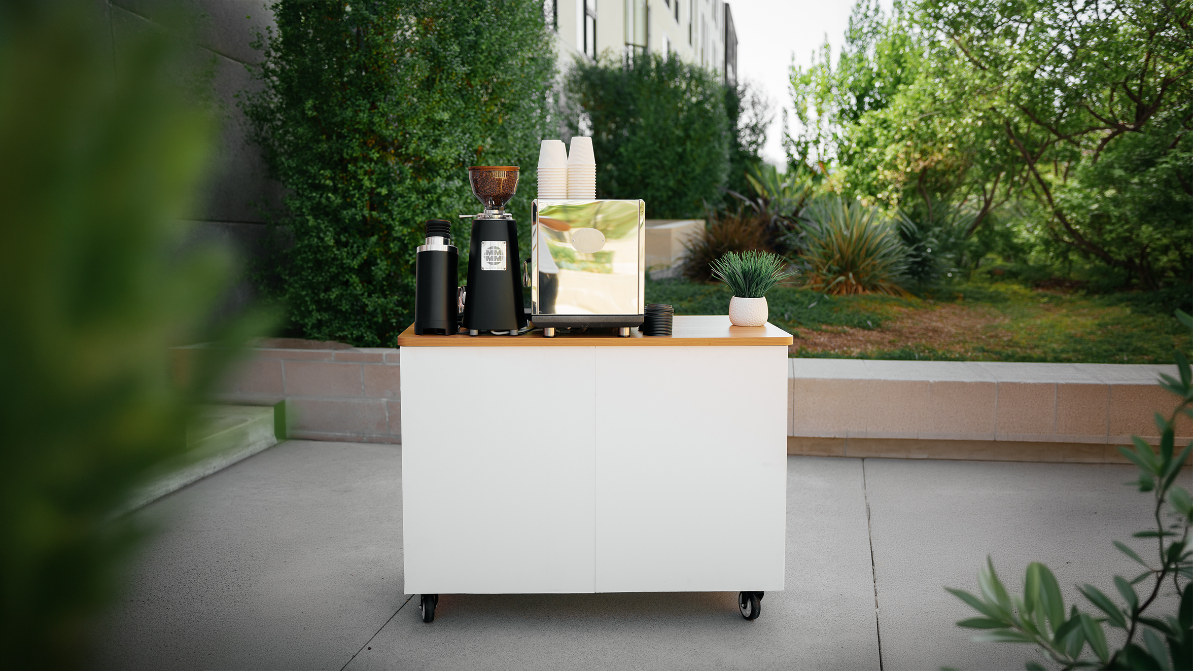 White mobile coffee cart with espresso machine, grinder, cups, and potted plant outdoors.
