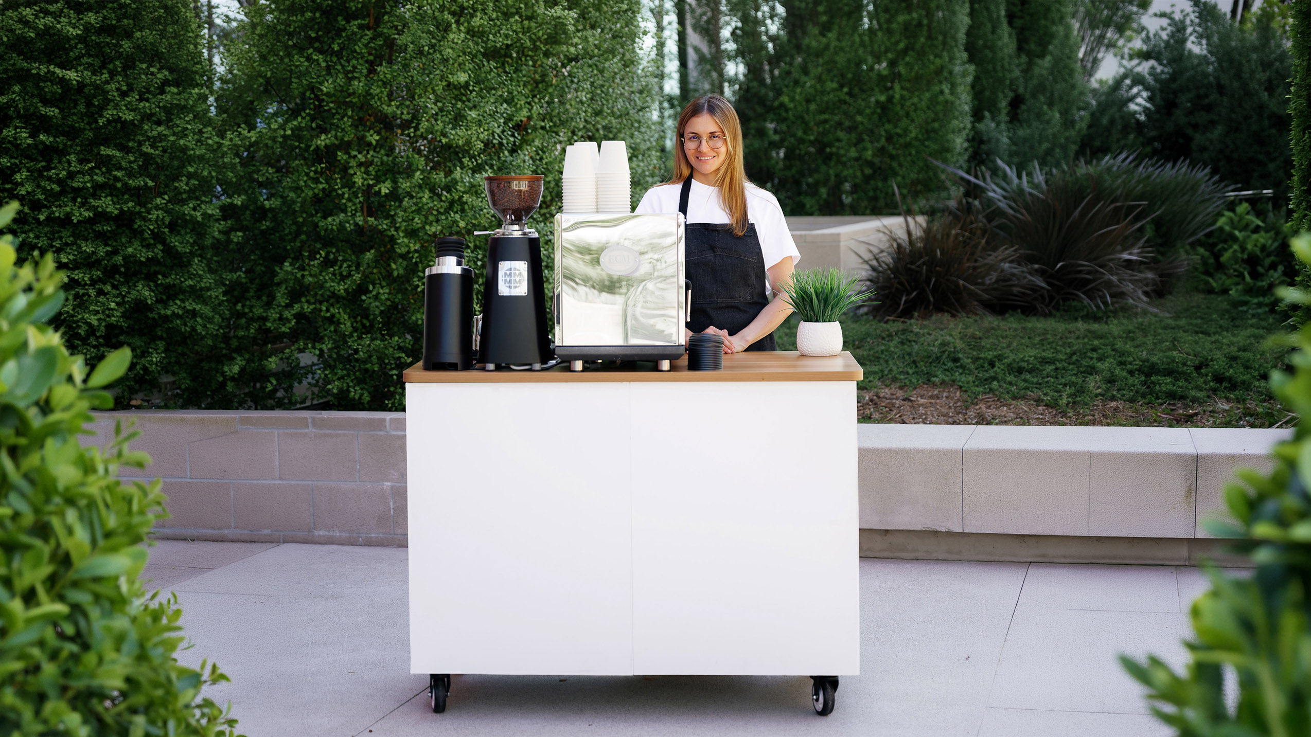 Woman with glasses and apron stands behind a mobile coffee cart outdoors with coffee equipment and plant.