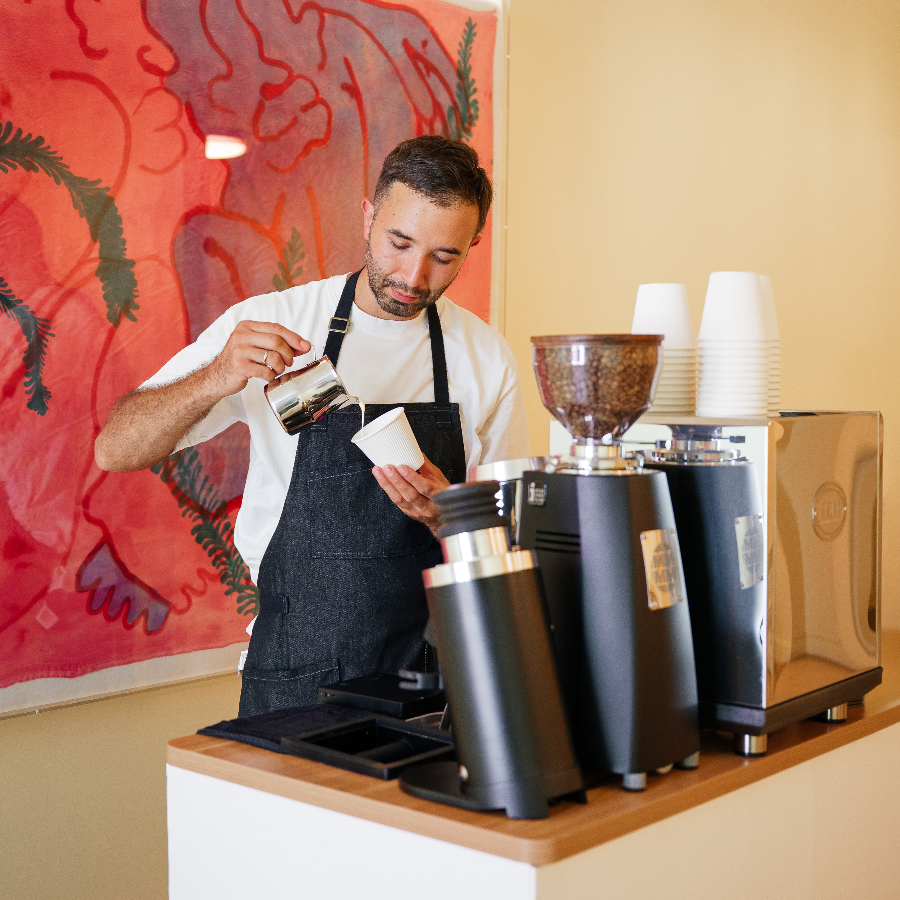 Barista pouring milk into a cup behind a coffee counter with coffee machines and cups.