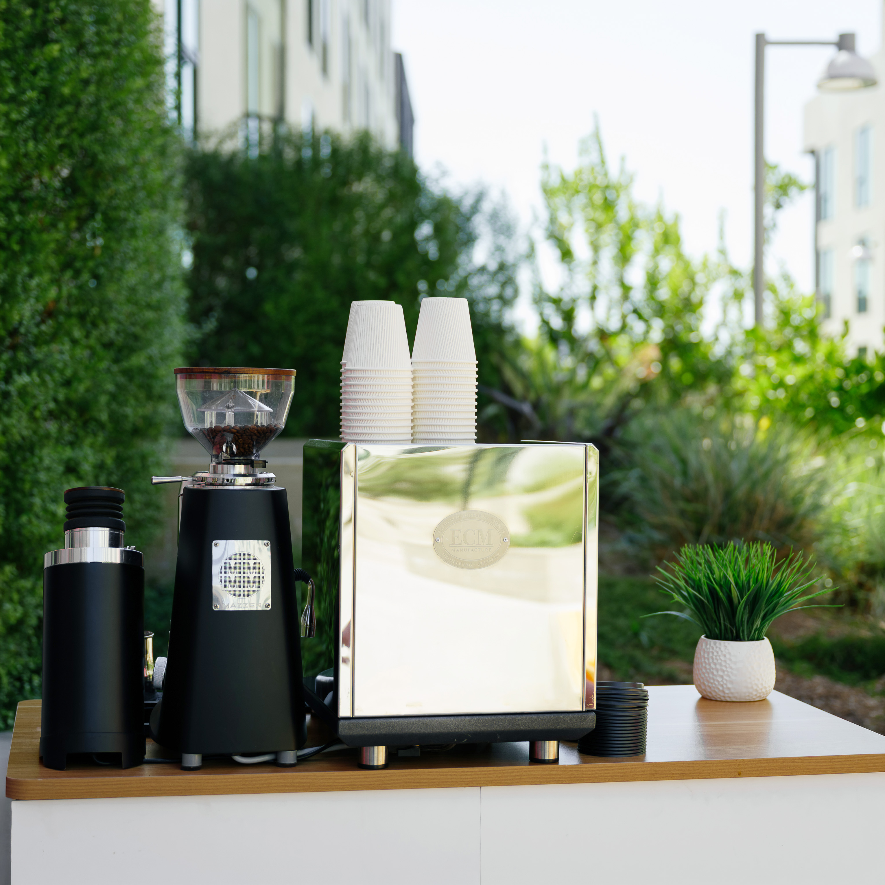 Coffee grinder and espresso machine with stacked paper cups on a wooden table outdoors.