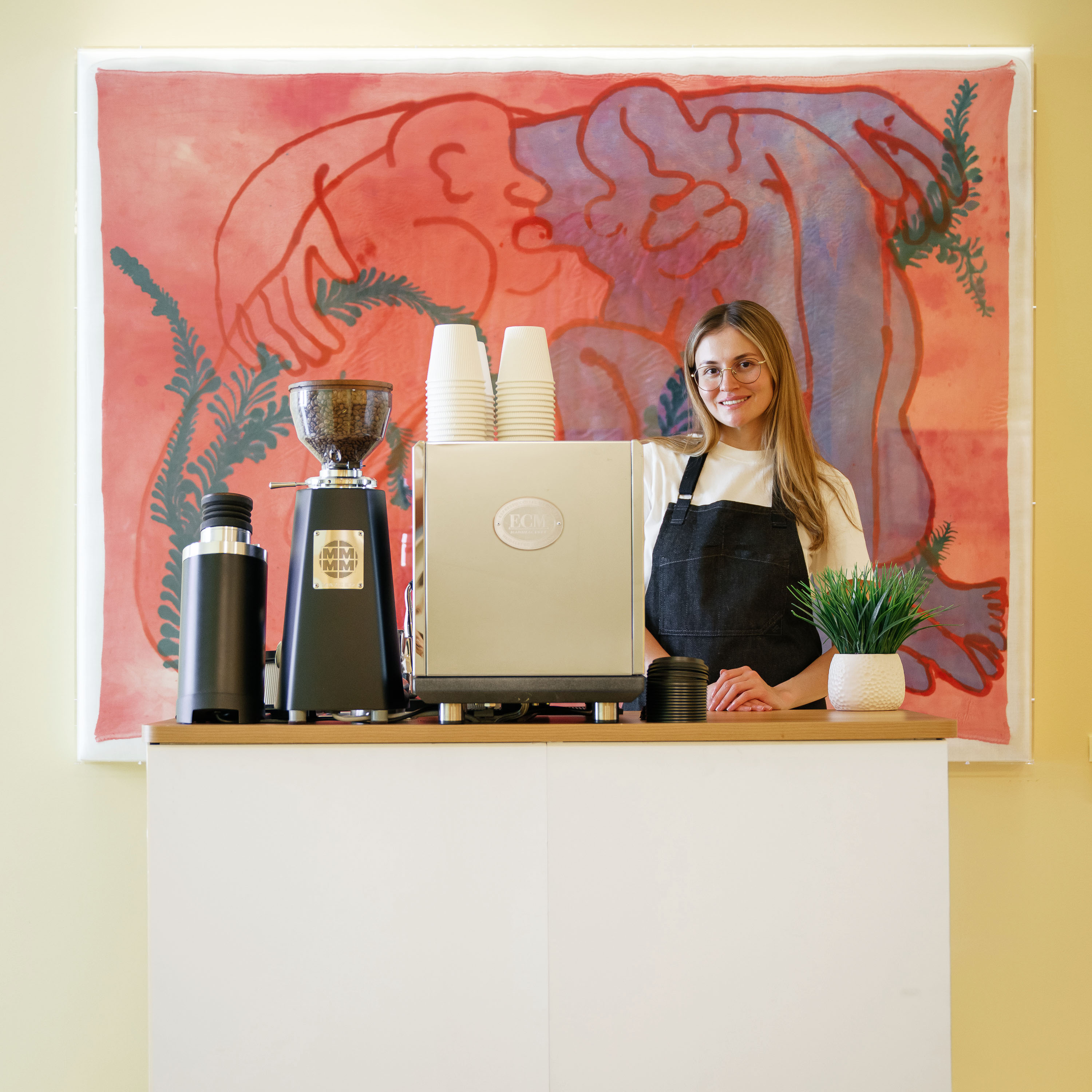 Smiling barista standing behind espresso machine and grinder at coffee counter with colorful backdrop.