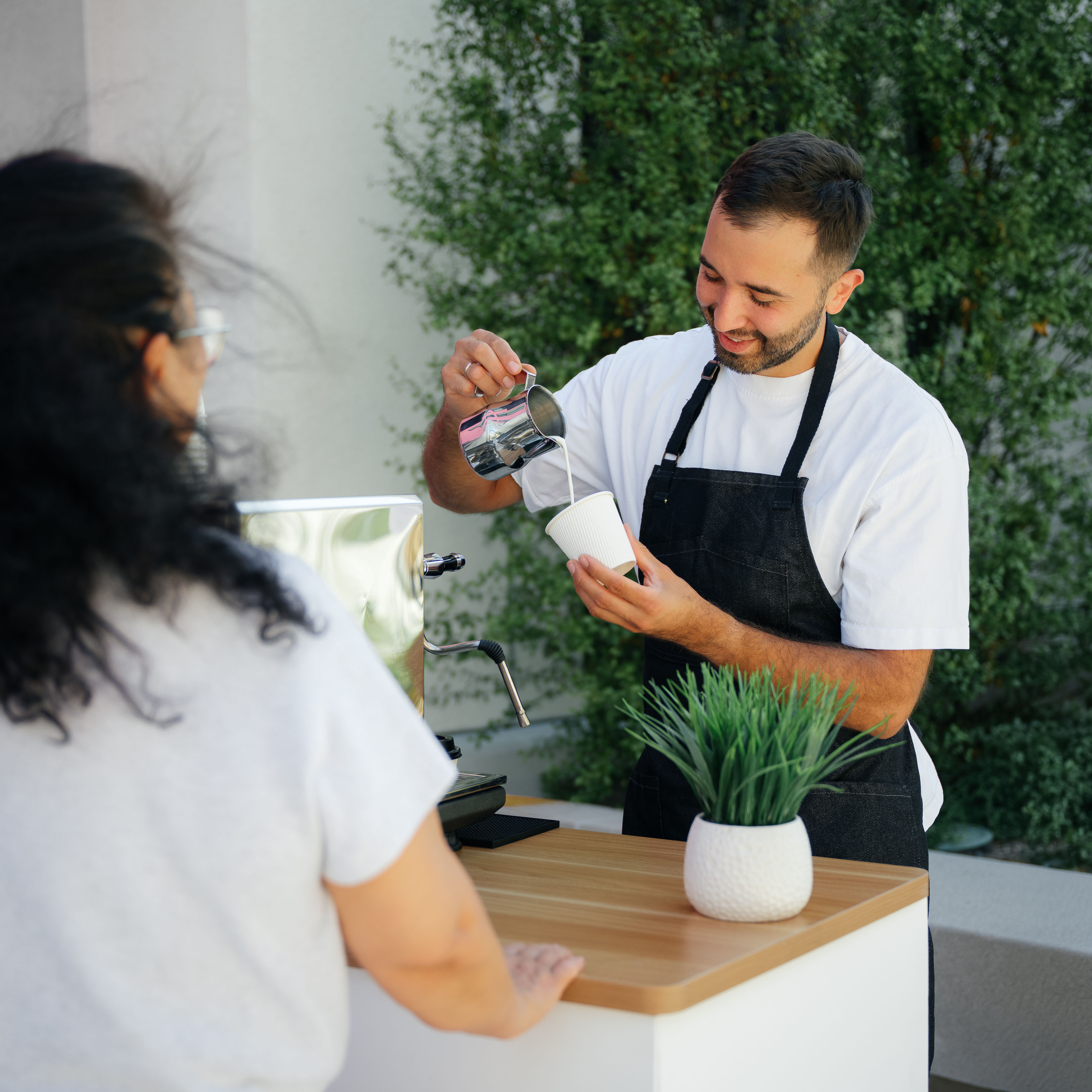Barista in apron pouring milk into a coffee cup at an outdoor counter with a customer.