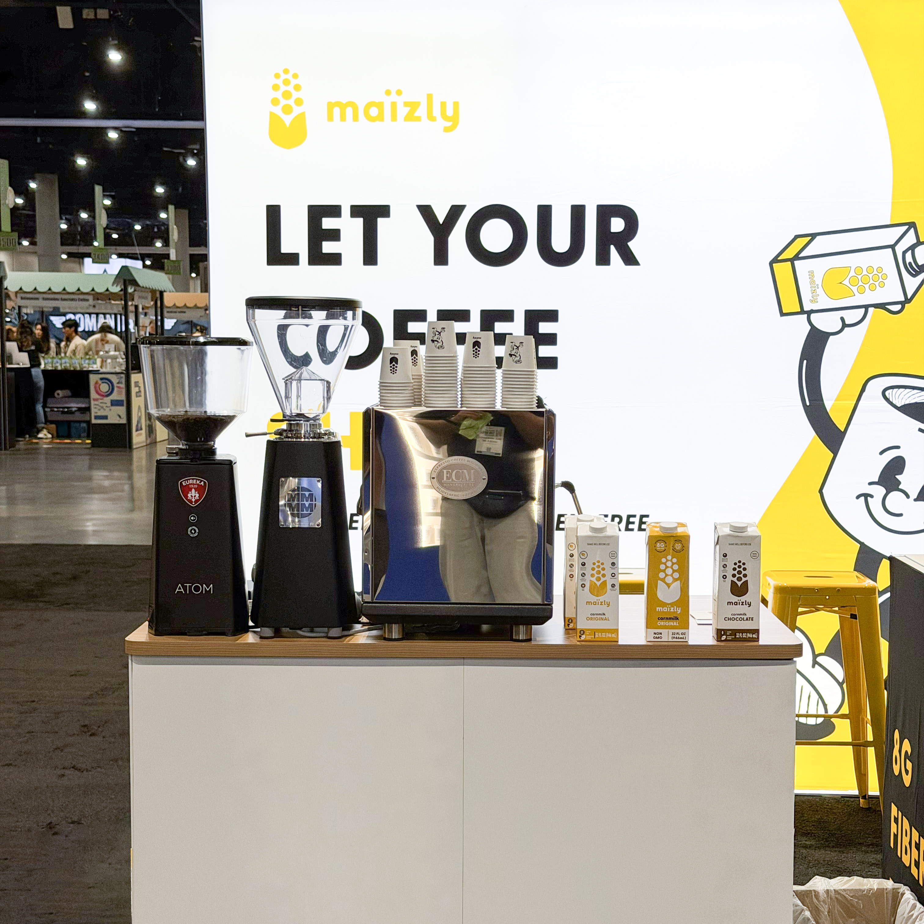 Coffee setup with grinders, espresso machine, and Maizly milk cartons on a counter at an event booth.