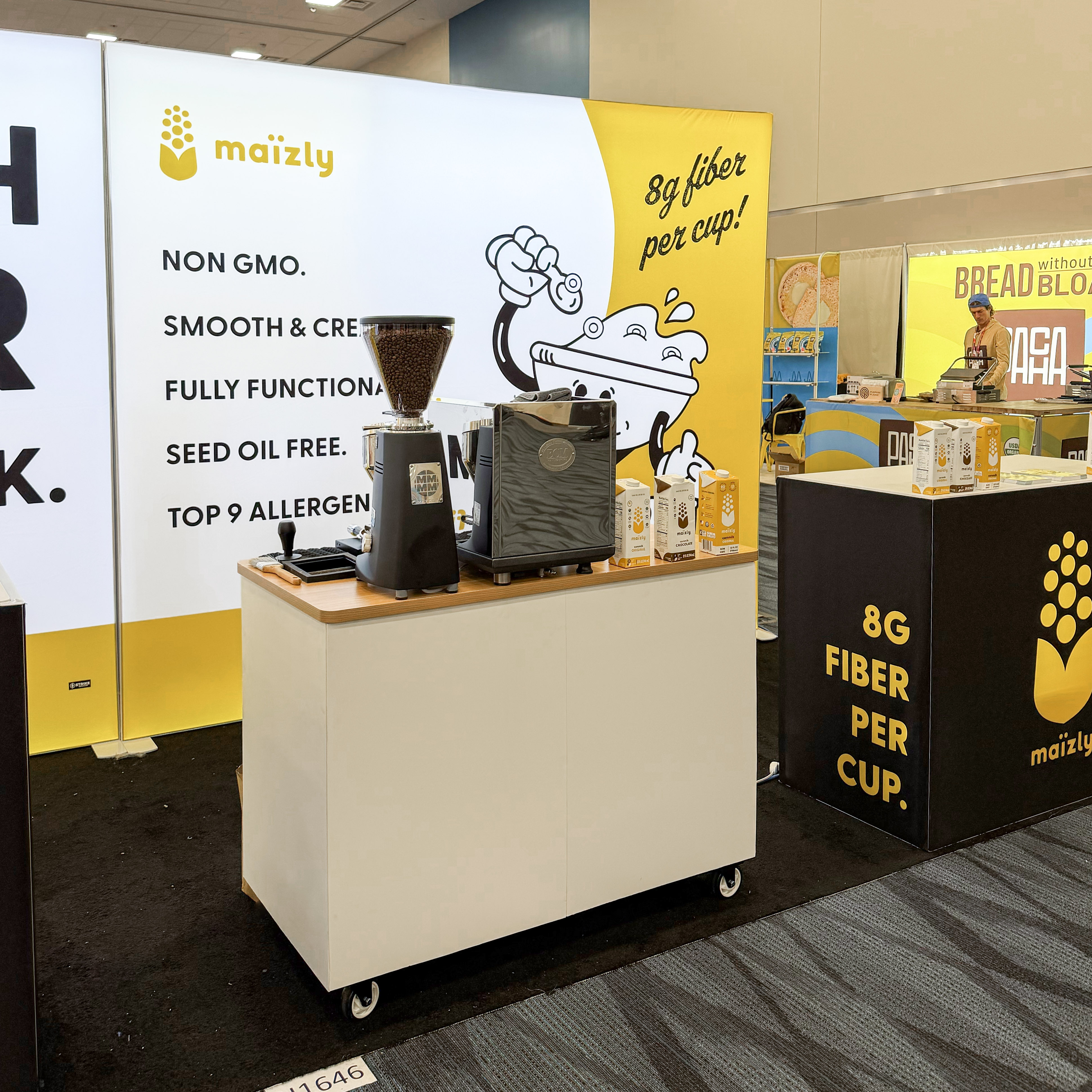 Maizly booth with coffee grinder, espresso machine, product boxes, and signage highlighting 8g fiber per cup.