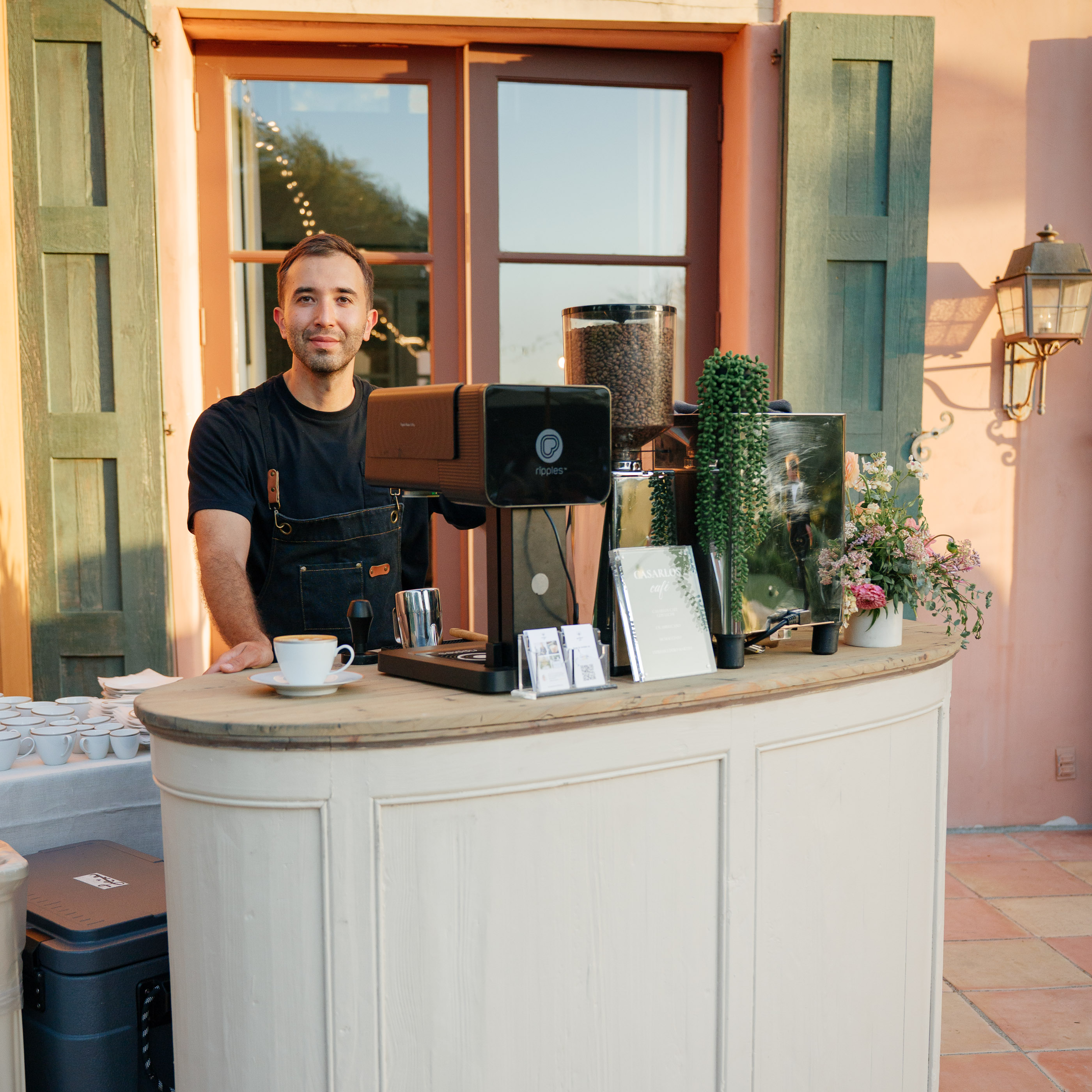 Barista standing behind a coffee machine at a rustic white curved counter with cups and flowers.