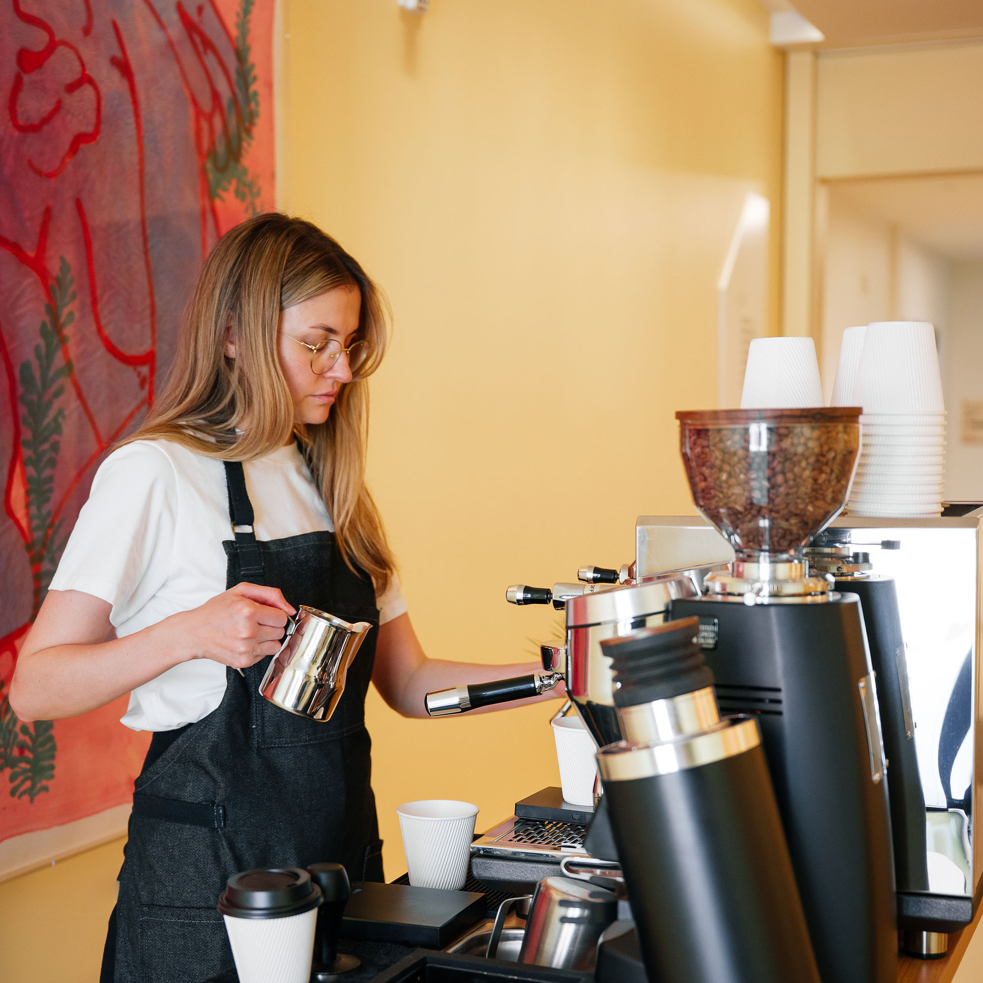 Woman barista with glasses and apron steaming milk using espresso machine in a coffee shop.