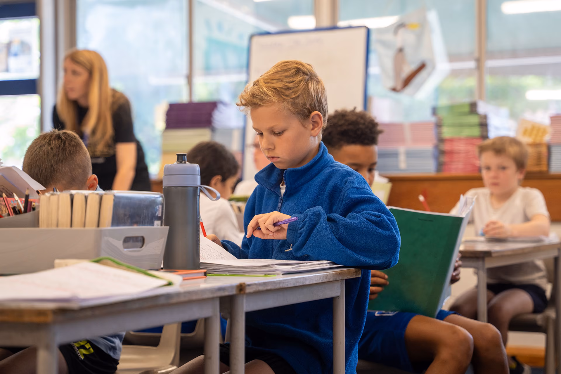 "Pupils working at desks in a busy classroom, with a teacher and bookshelves in the background.