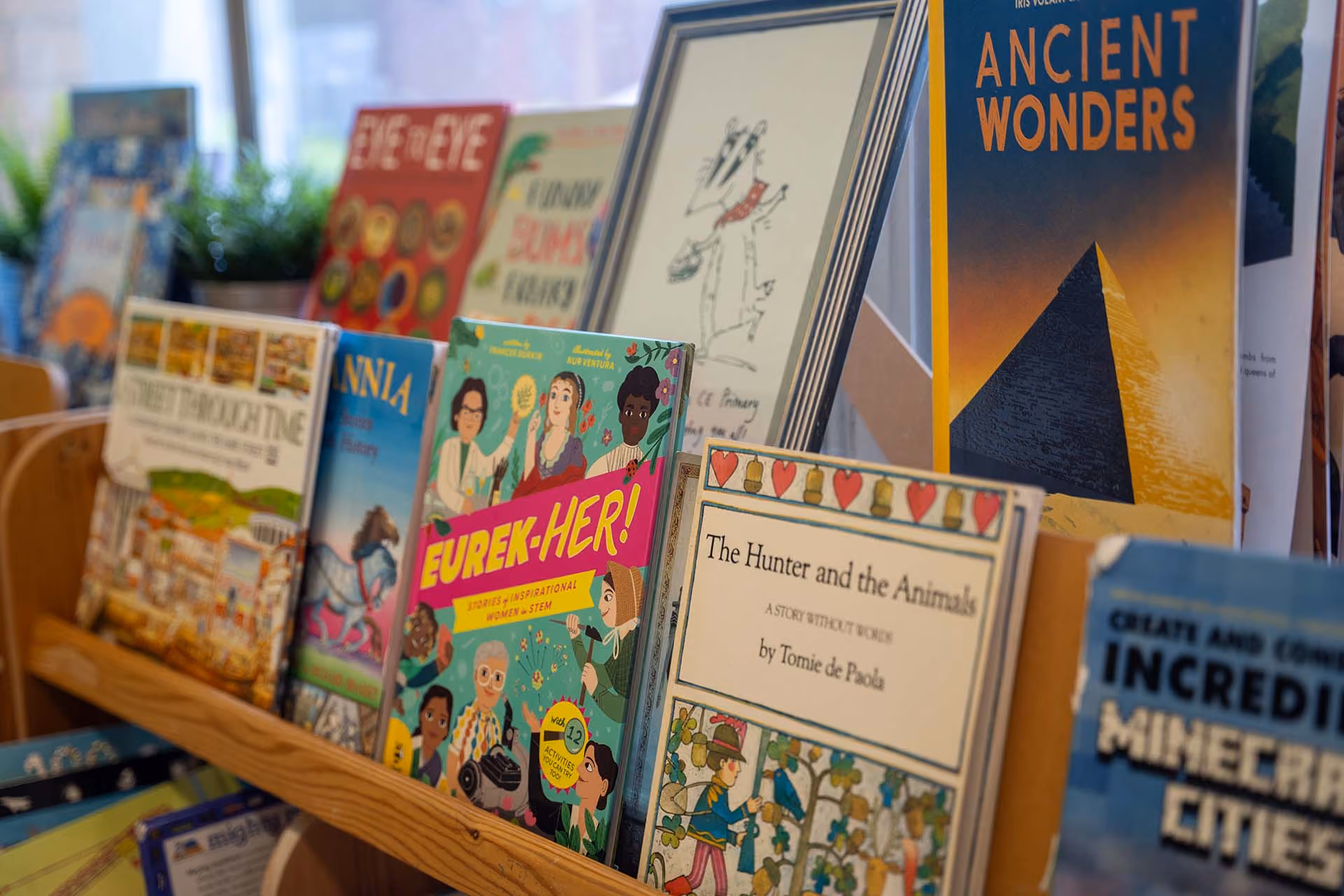 Row of children's books on a classroom bookshelf, including titles like 'Eurek-Her!' and 'Ancient Wonders'.