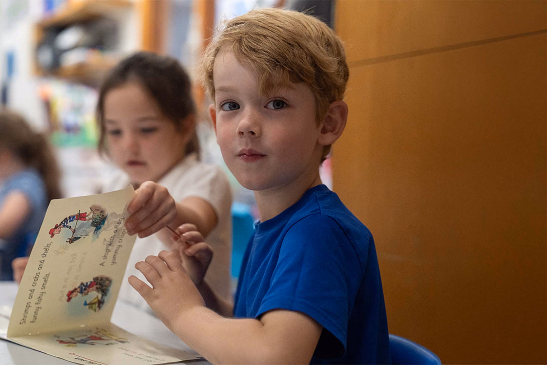 Young pupil in a blue school t-shirt looking towards the camera while writing at a classroom table.