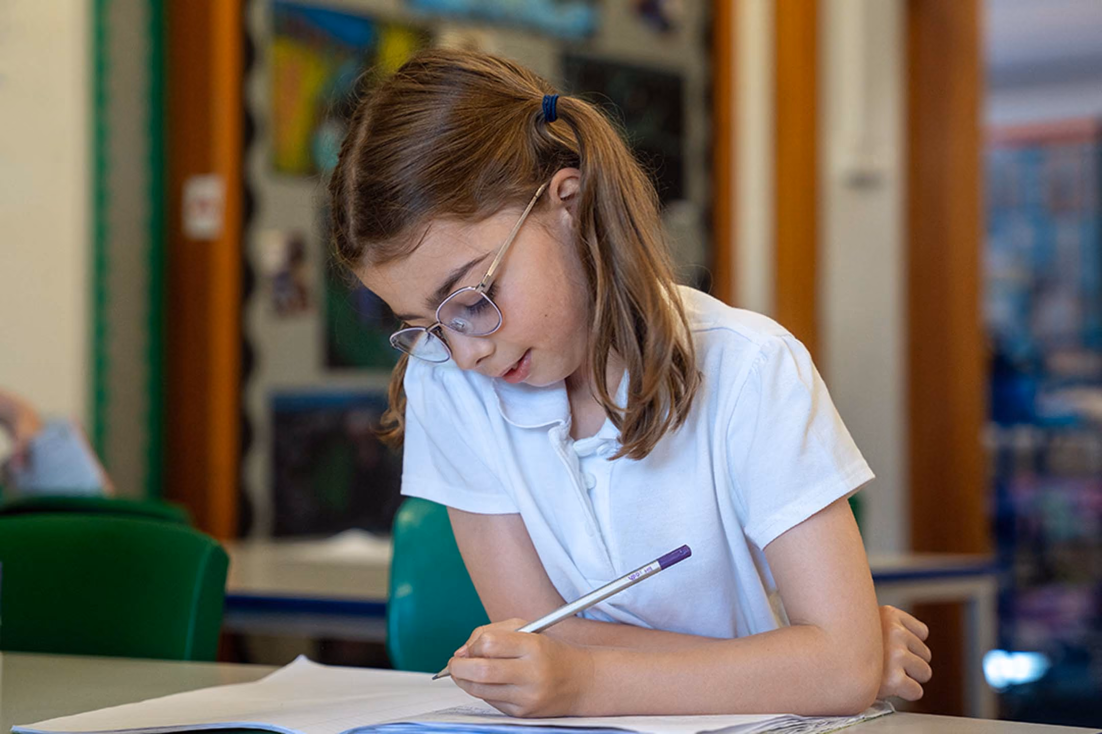Pupil in glasses and white school polo shirt writing with a pencil at her desk.