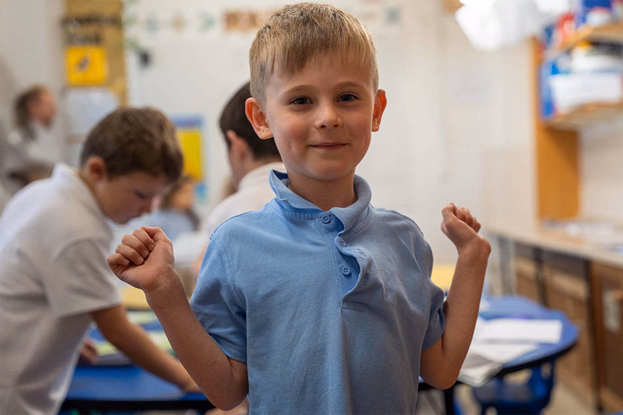 Pupil in a light blue polo shirt smiling with raised fists in a classroom.