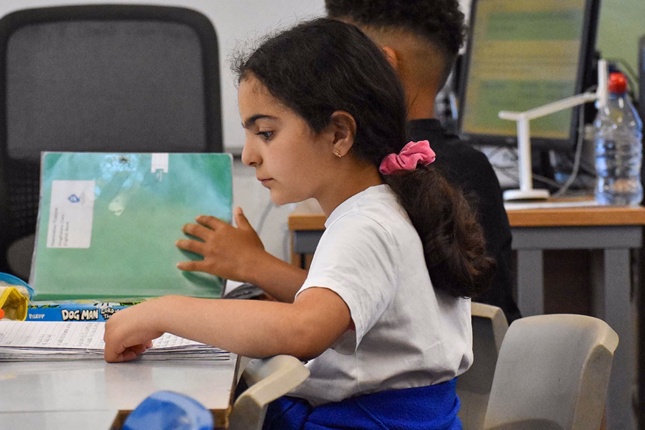 Pupil at a classroom desk reading from a book, with a computer and other pupils in the background.