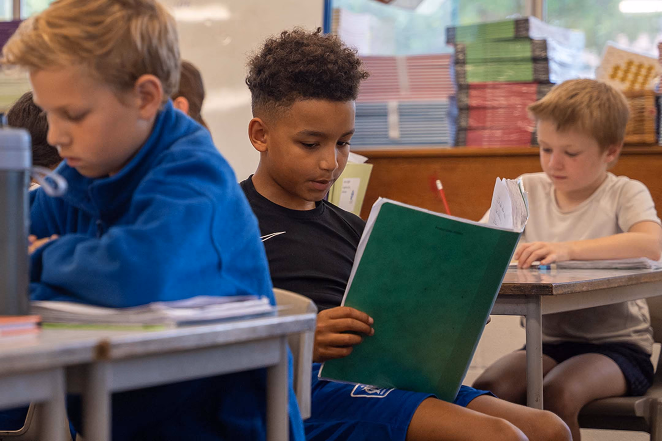 Pupils reading and writing at desks in a classroom, with books stacked in the background.