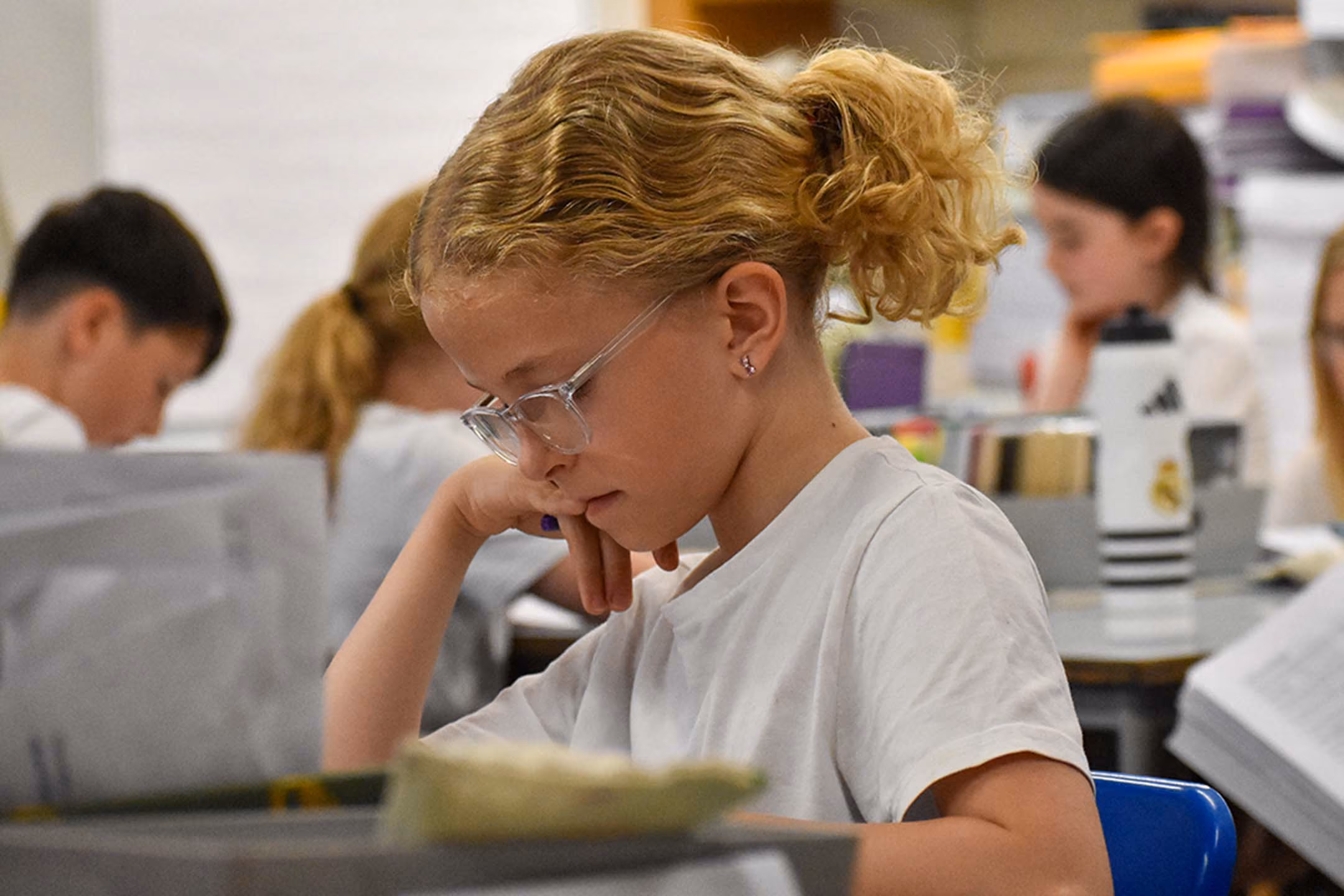Pupil in glasses concentrating on work at a classroom desk, with other pupils in the background.