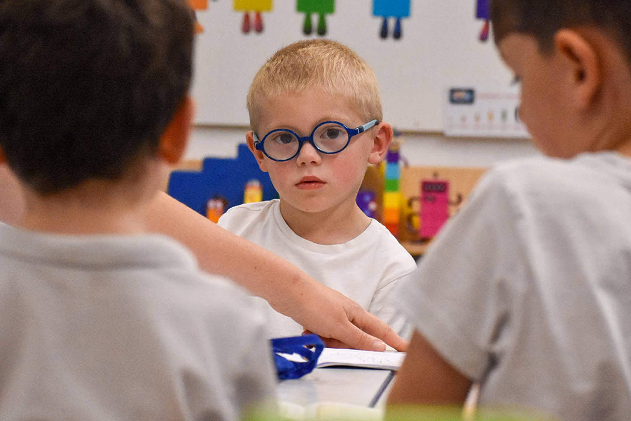 Young pupil in blue glasses sitting at a classroom table with two other pupils, colourful robot decorations on the wall behind.