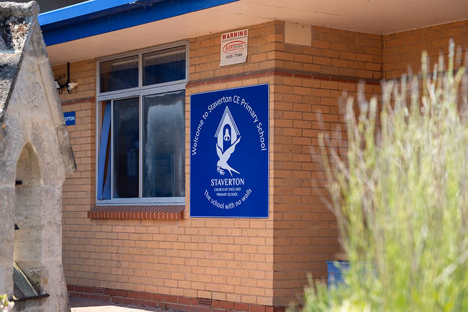 Blue welcome sign for Staverton Church of England Primary School on a brick building.