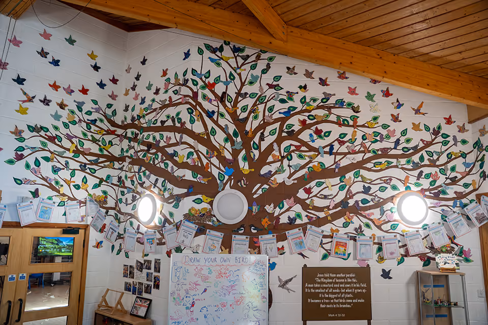 Wall mural of a tree with colourful paper birds and leaves, a whiteboard, and a Bible quote.