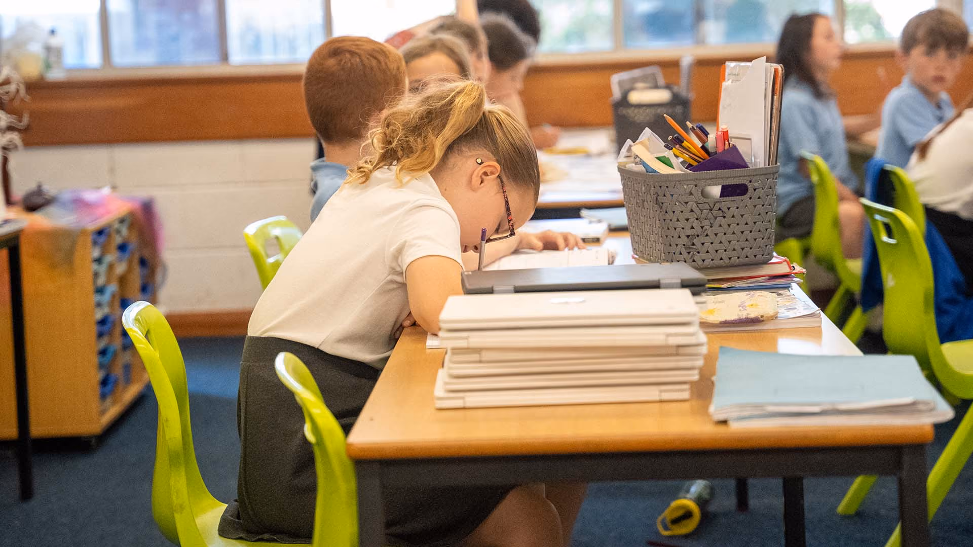 Pupil in glasses leaning over her desk to write, beside a stack of closed laptops.