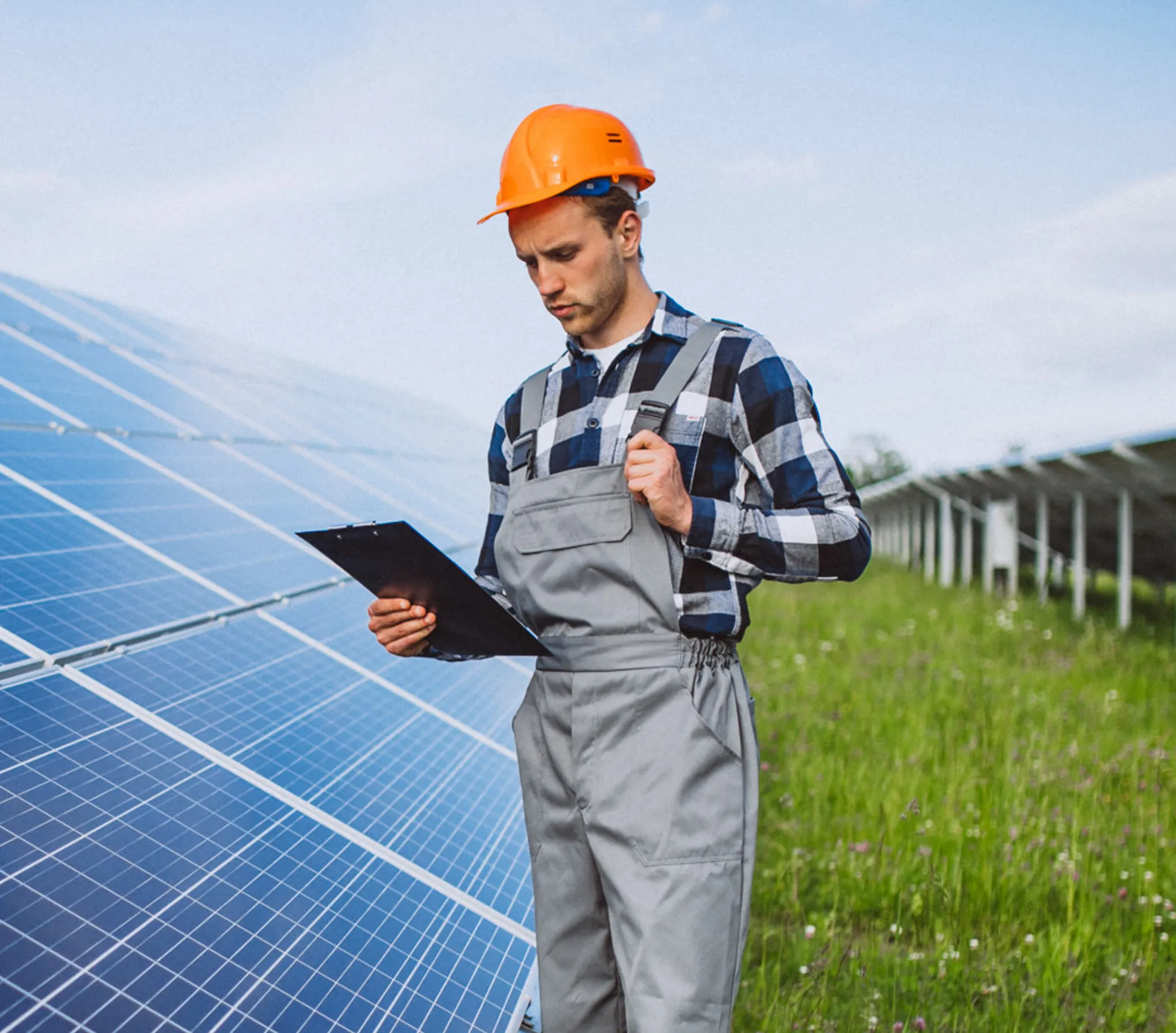 Engineer in orange helmet and overalls checking clipboard near large solar panels outdoors.