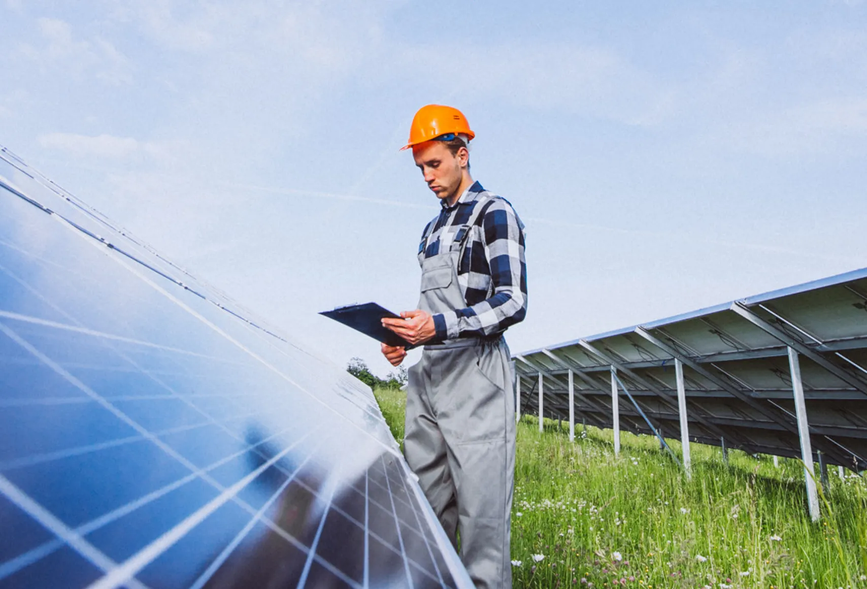 Engineer in gray overalls and orange helmet inspecting solar panels while holding a clipboard in a grassy field.