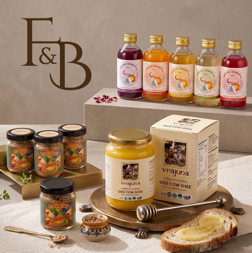 Display of food and beverage products featuring five bottles of colorful bottled drinks labeled "Drinking Kombucha" in various flavors with gold caps on a raised platform; several small jars of Rajhing spice mix with black lids on a golden tray; a jar and box of Vrajuna Desi Cow Ghee on a brass plate with a honey dipper; a slice of bread spread with ghee; all arranged on a beige tablecloth with a beige background and brown F&B logo on the left.