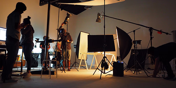 Dimly lit photo studio with five people setting up cameras and lighting equipment around a blank whiteboard and a large monitor on easels.