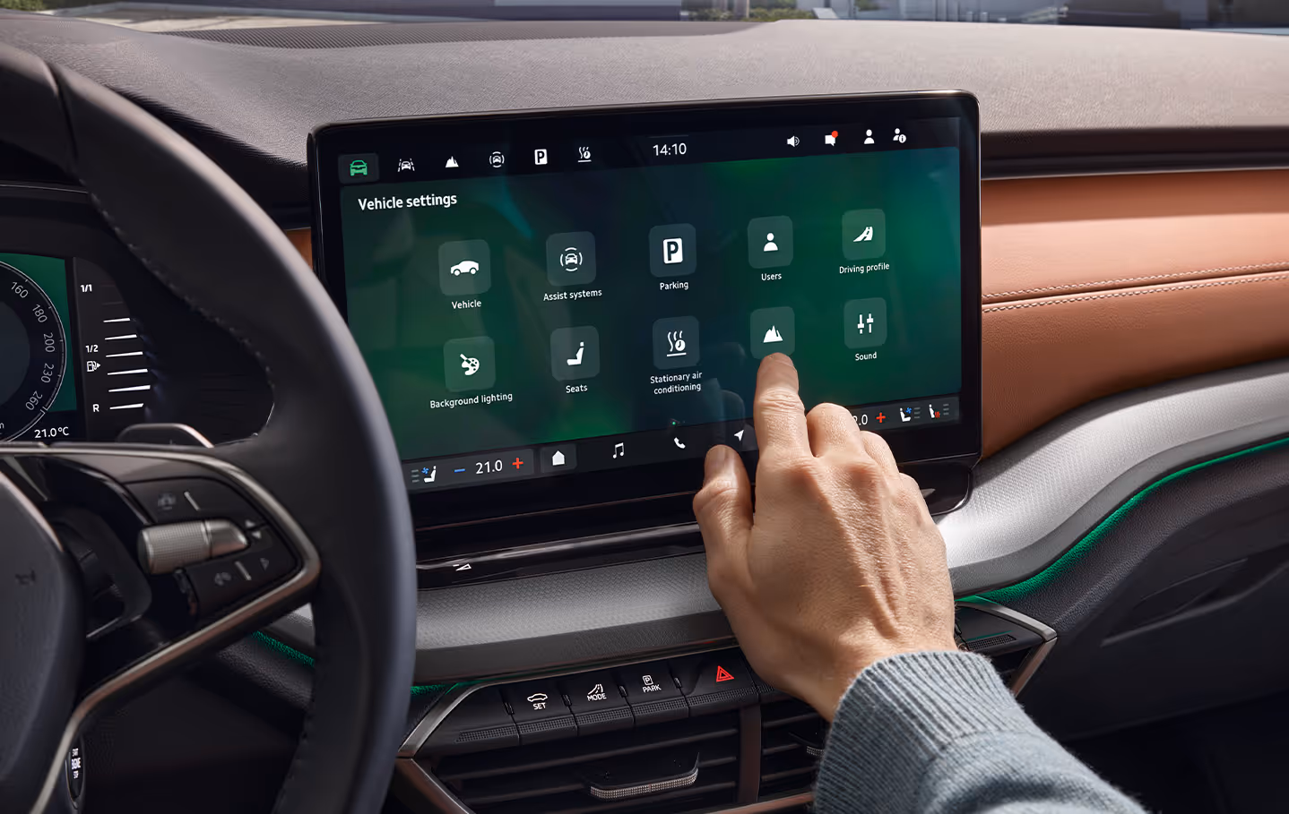 Nahaufnahme des Infotainment-Displays im Interieur des Škoda Octavia 30 Jahre. Eine Hand bedient den großen Touchscreen, auf dem das Menü für Fahrzeugeinstellungen wie Assistenzsysteme, Sitze und Klimatisierung zu sehen ist. Der Bildausschnitt zeigt zudem Details des Multifunktionslenkrads sowie die grüne Ambientebeleuchtung unterhalb des Armaturenbretts mit braunem Bezug.