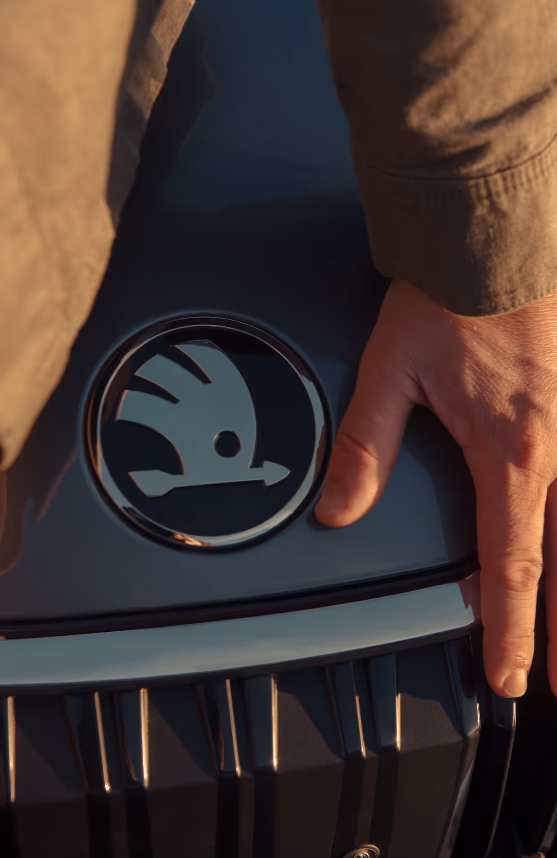 Eine Nahaufnahme zeigt die Hand eines Mannes, die sanft auf der Motorhaube des Škoda Octavia 30 Jahre ruht, direkt neben dem markanten Škoda Logo. Das Bild fokussiert auf die Design-Details der Frontpartie und den Kühlergrill, die von warmem, goldenem Sonnenlicht angestrahlt werden.