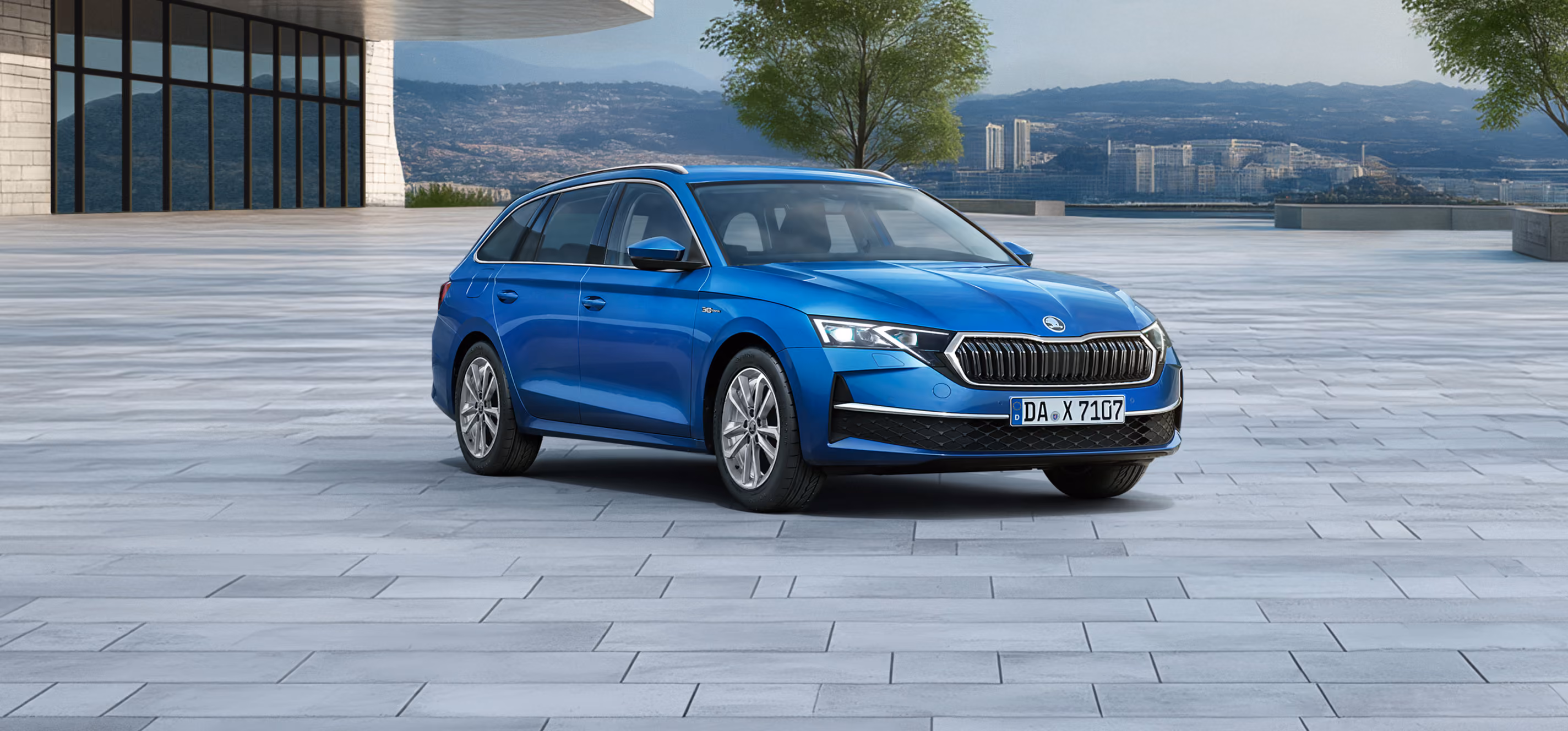 Das Bild zeigt das Sondermodell Škoda Octavia Combi 30 Jahre in der Farbe Race-Blau Metallic aus der vorderen linken schrägen Perspektive auf einem weitläufigen, gepflasterten Platz vor einer Stadtkulisse. Zu sehen sind Exterieur-Details wie die silbernen Leichtmetallfelgen, der Kühlergrill mit Chromrahmen sowie die schwarze Dachreling. Am unteren linken Bildrand hebt ein hellgrüner Störer einen Preisvorteil sowie die inkludierte 5-Jahres-Garantie hervor.