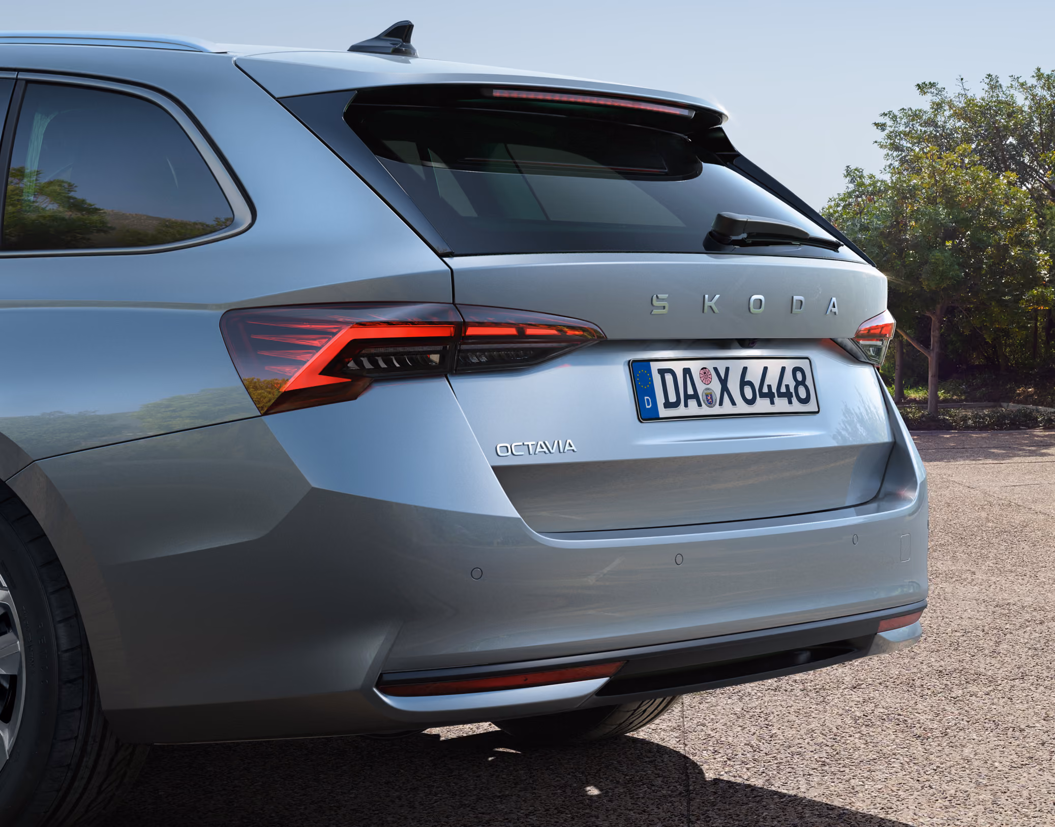 Seitliche Rückansicht eines Škoda Octavia Combi 30 Jahre. Im Fokus stehen die markanten LED-Heckleuchten, der mittig platzierte Škoda Schriftzug auf der Heckklappe sowie die Leichtmetallfelgen. Das Fahrzeug steht bei Tageslicht auf einem gepflasterten Untergrund vor einer Reihe grüner Bäume.