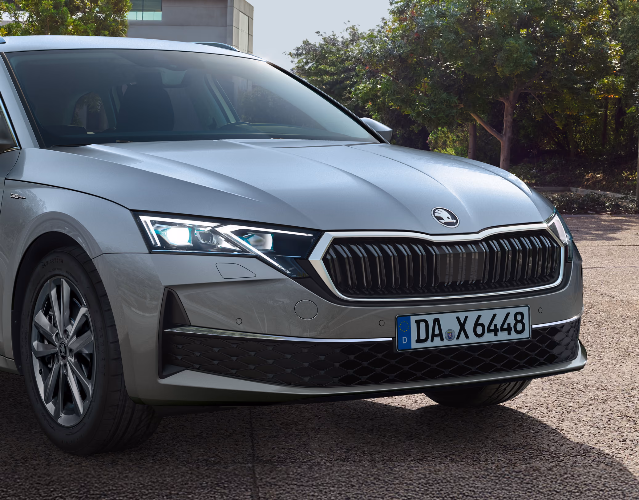 Seitliche Nahaufnahme der Frontpartie eines Škoda Octavia 30 Jahre. Im Fokus stehen der markante Matrix-LED-Scheinwerfer auf der Beifahrerseite, der Kühlergrill mit Chromrahmen sowie die glanzgedrehten Leichtmetallfelgen. Das Fahrzeug befindet sich auf einem gepflasterten Weg vor einer Kulisse aus grünen Bäumen und hellem Tageslicht.