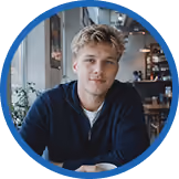 Junger Mann mit blondem Haar sitzt entspannt in einem Café, lächelt in die Kamera, moderne Inneneinrichtung im Hintergrund.