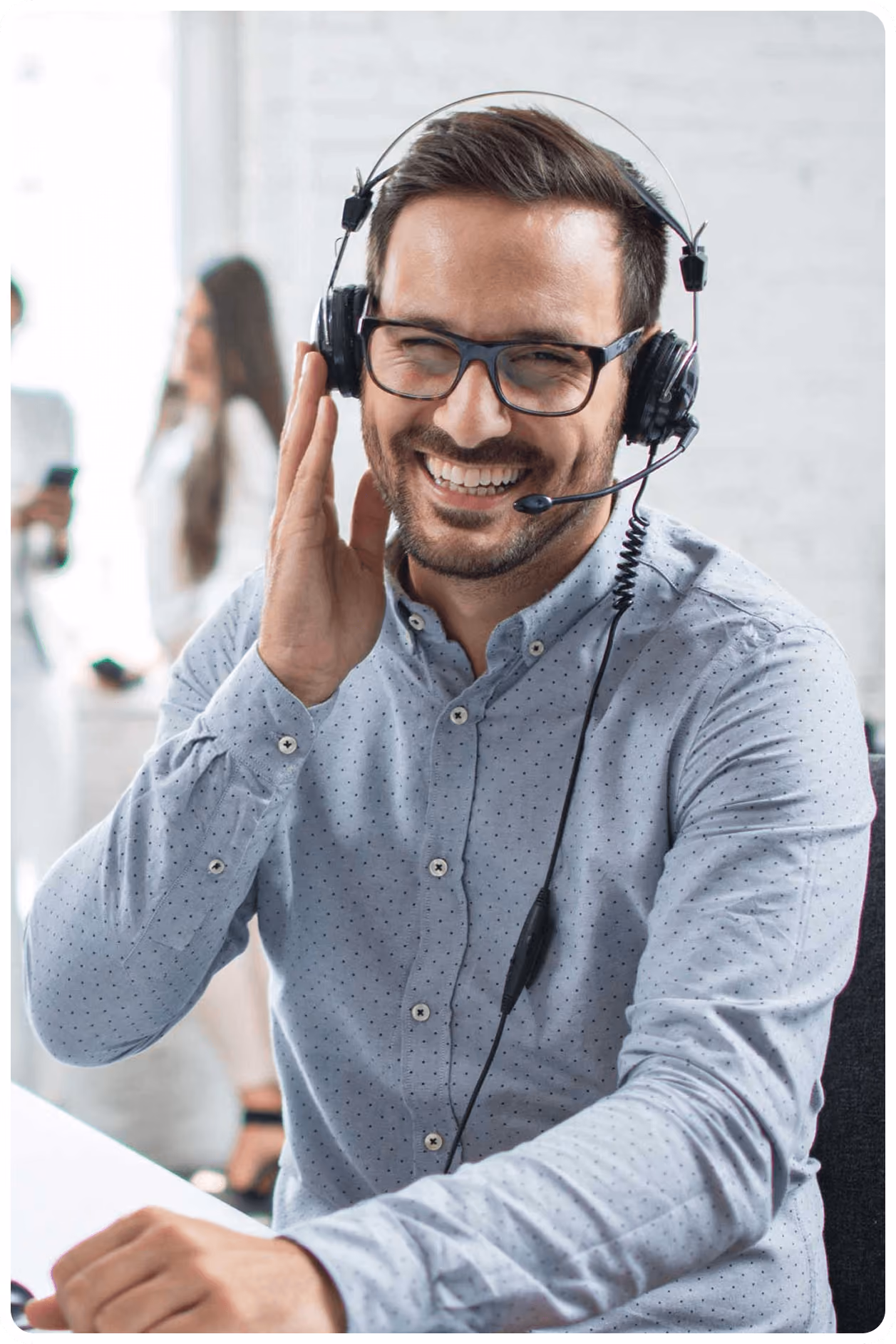 Lächelnder Mann mit Brille trägt Headset, sitzt im Büro, im Hintergrund Kollegen verschwommen sichtbar.