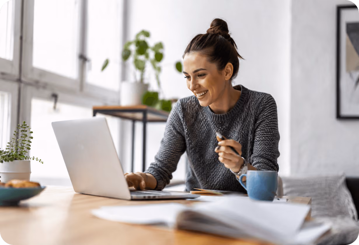Frau arbeitet am Laptop, lächelnd, mit Stift; helle Wohnung, Pflanzen im Hintergrund.