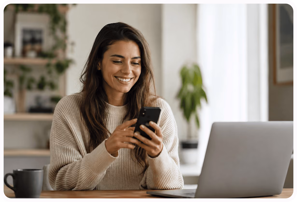 Frau lächelnd, nutzt Smartphone am Tisch mit Laptop, Pflanzen im Hintergrund.