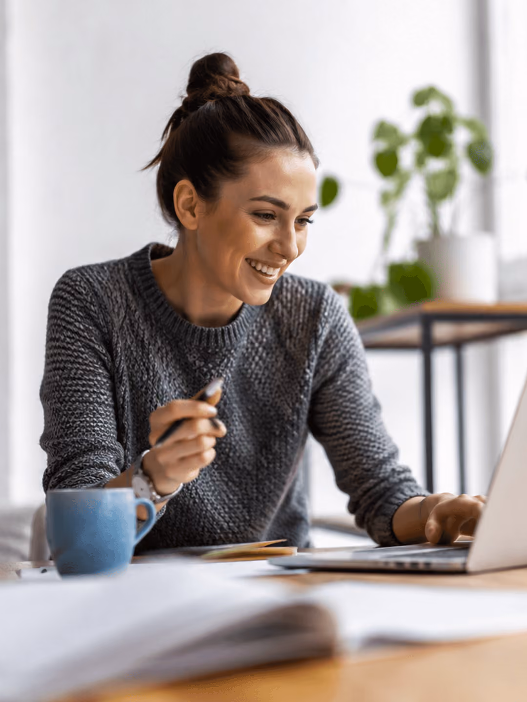 Frau arbeitet lächelnd an einem Laptop, hält Stift in der Hand, Pflanze im Hintergrund, blauer Becher auf dem Tisch.