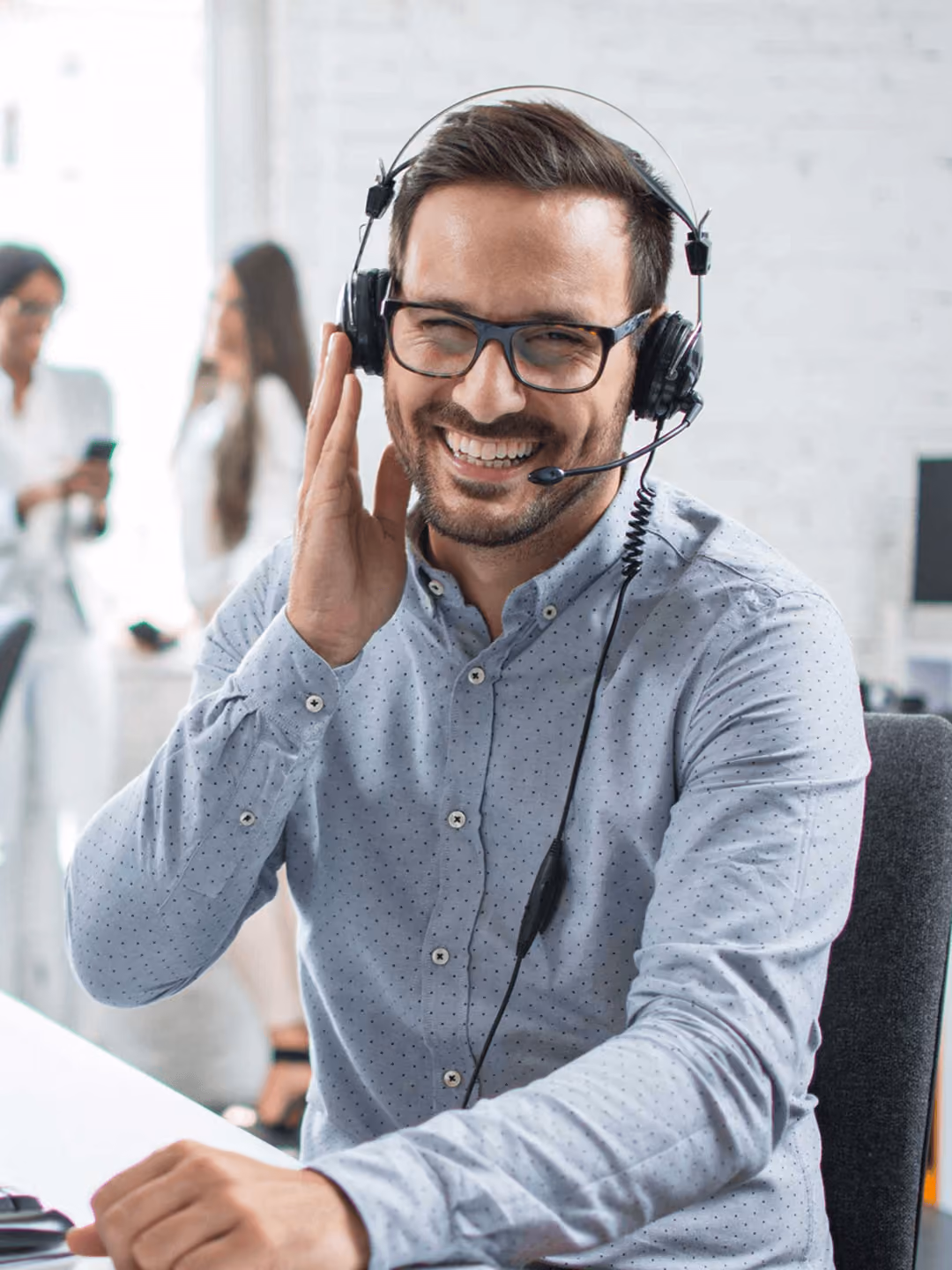 Lächelnder Mann mit Headset in modernem Büro, telefoniert und trägt eine helle, gepunktete Bluse. Kolleginnen im Hintergrund.
